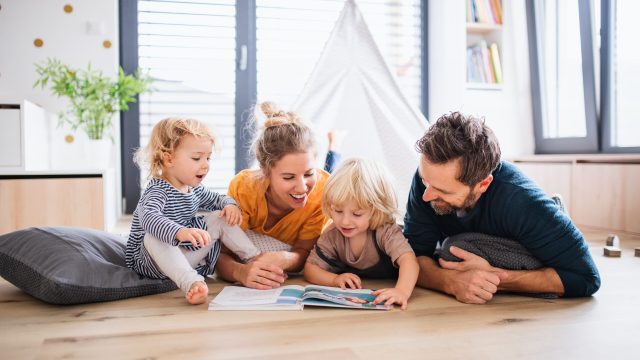 Front view of young family with two small children indoors in bedroom reading a book.